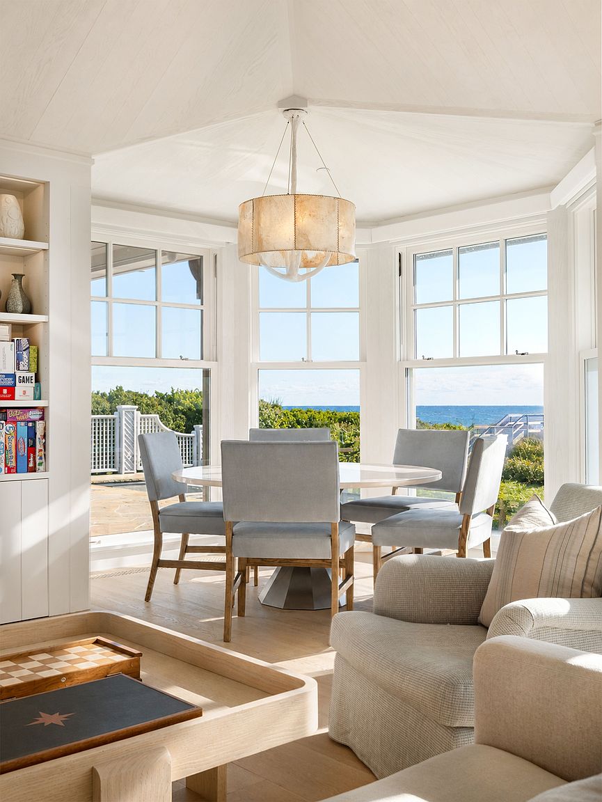 This bright and airy dining area features a round table surrounded by four light-gray upholstered chairs, positioned in a bay window nook that offers expansive ocean views. To the left, a built-in shelving unit displays various games and decor, while a textured drum pendant light hangs above the table. The foreground includes a portion of a light wood coffee table and a comfortable armchair, creating a cohesive and inviting open-concept living space.