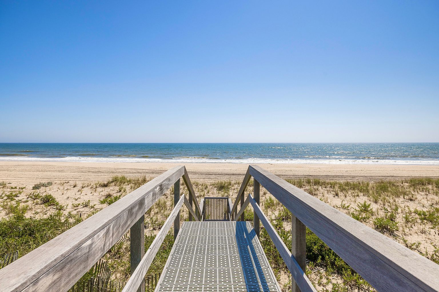 This image captures a direct, eye-level perspective of a wooden boardwalk leading down to a pristine, sandy beach. The walkway features sturdy railings and a metal grate surface, framed by natural dune grasses under a clear, bright blue sky. The composition emphasizes a direct path to the ocean, highlighting the property's convenient and scenic beach access.