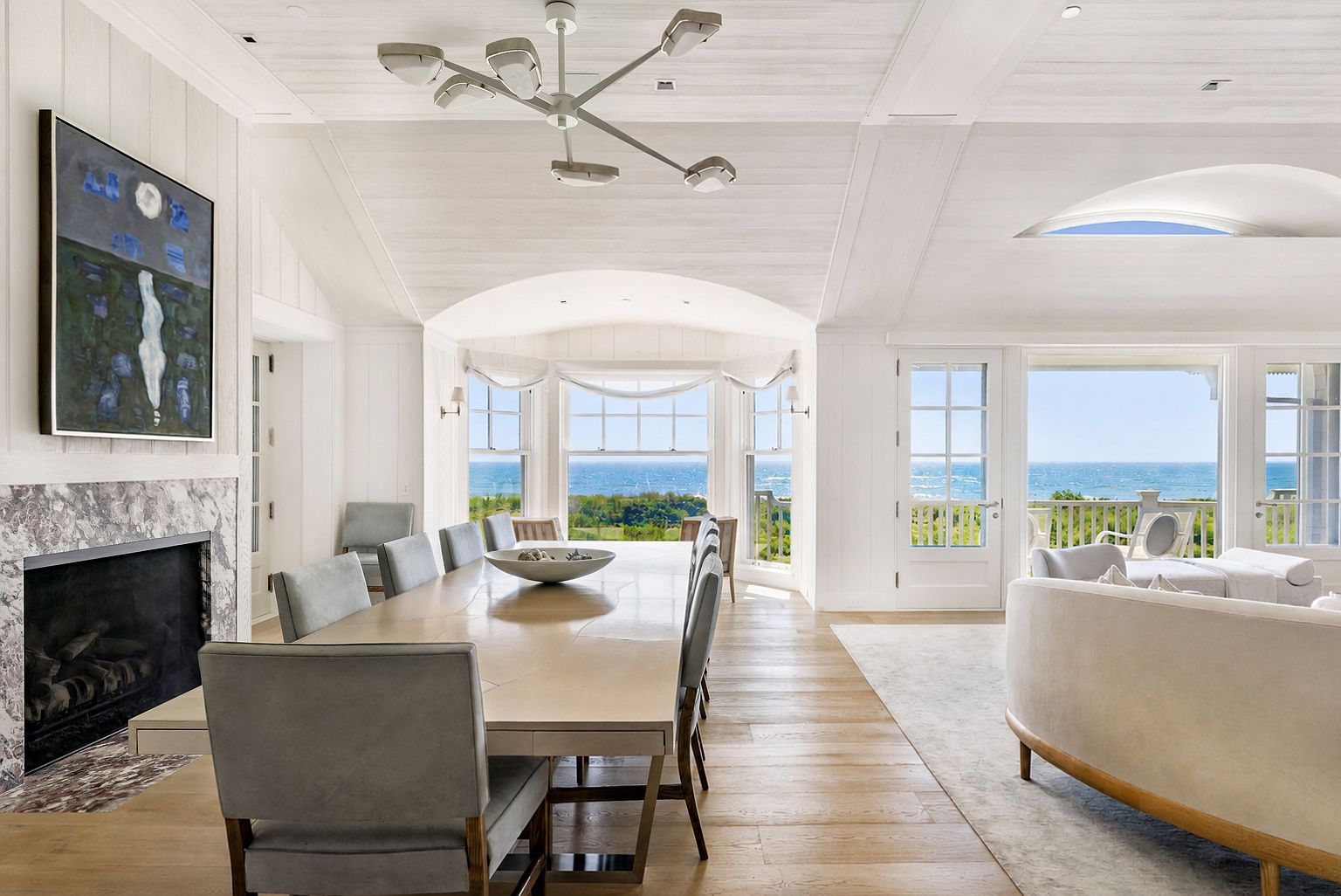 This bright, open-concept dining area features a long table surrounded by upholstered chairs, positioned beneath a modern, multi-armed chandelier. To the left, a stone-clad chimney breast houses a fireplace, while the right side opens into a spacious living area with a curved sofa. Large windows and glass doors provide expansive, unobstructed views of the ocean, creating a serene and airy atmosphere.