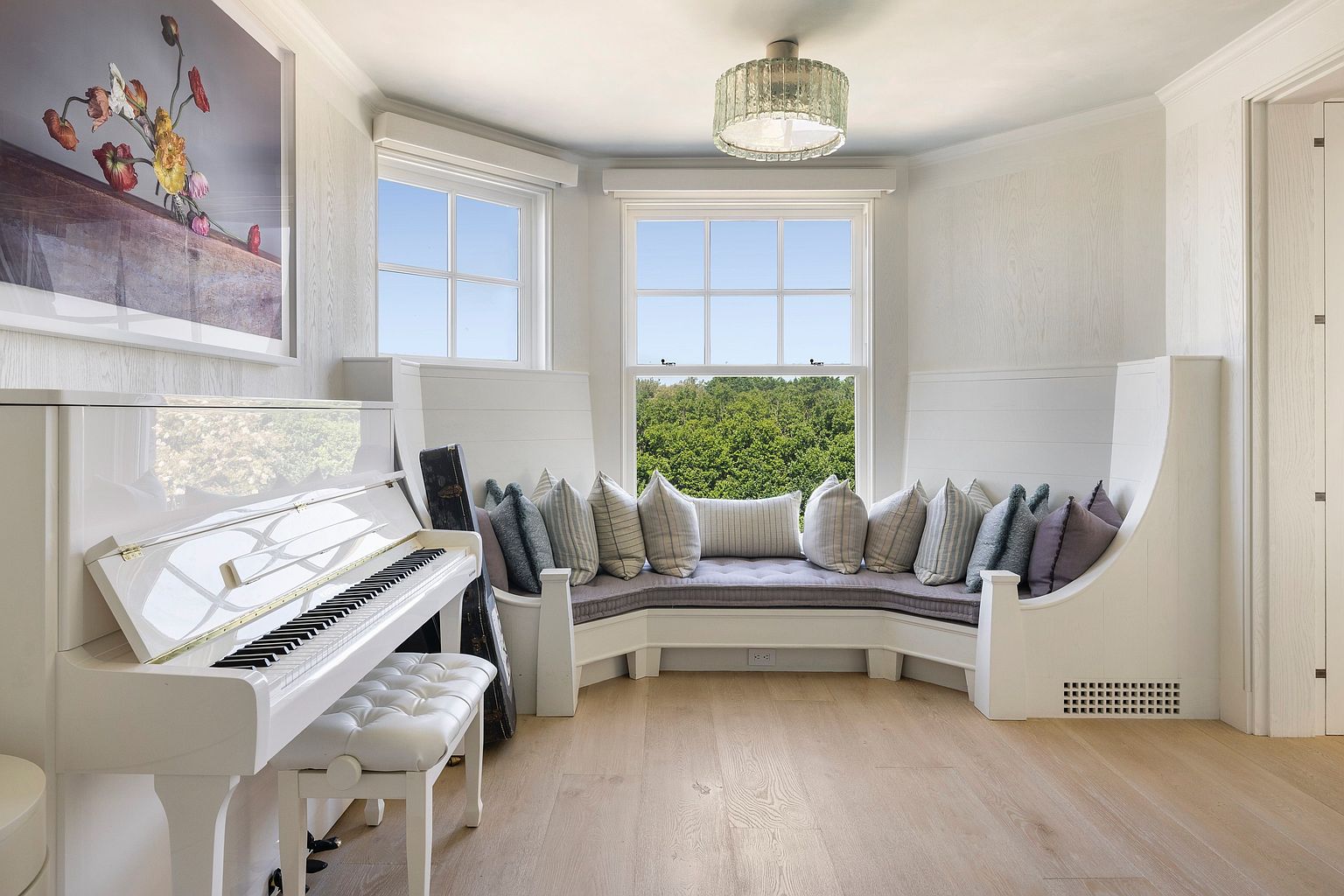 This bright and airy room features a white piano positioned next to a large, curved built-in window seat lined with plush, patterned cushions. The space is finished with light wood flooring and white paneled walls, creating a serene and elegant atmosphere. A crystal-style chandelier hangs from the ceiling, while a large floral art piece adds a touch of color to the otherwise neutral palette.