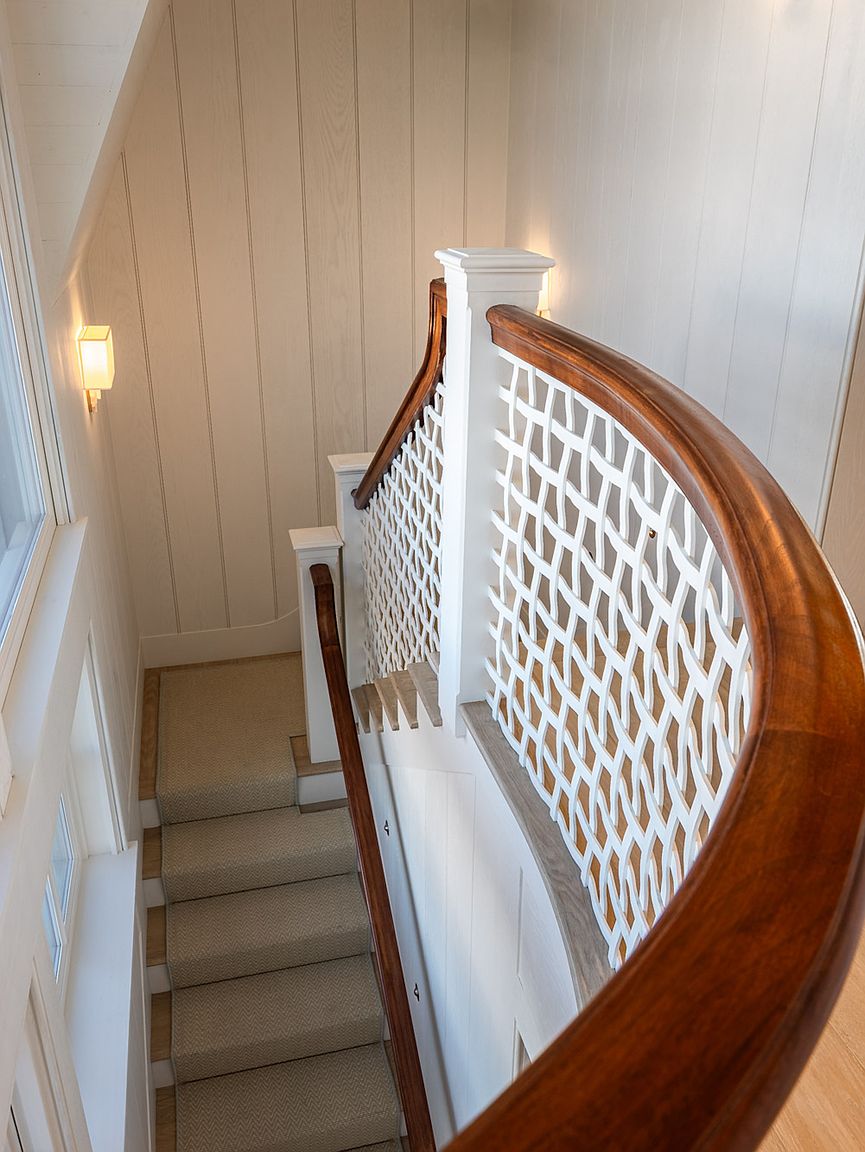 This high-angle perspective captures a graceful, curved staircase featuring a rich, dark wood handrail and a unique white lattice balustrade. The stairs are carpeted in a neutral tone, leading down past light-colored vertical wall paneling and a wall-mounted sconce. The design emphasizes architectural elegance and a bright, airy aesthetic.