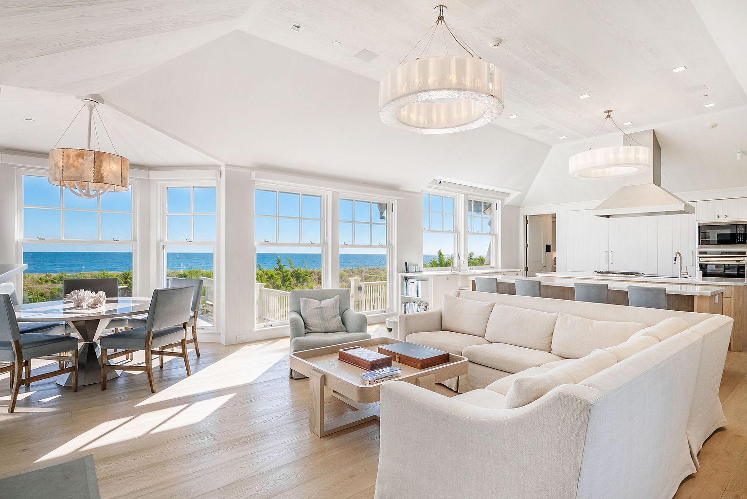 This bright, open-concept living and dining area features a coastal aesthetic with light wood flooring and vaulted ceilings. A large, plush sectional sofa and a central coffee table anchor the living space, while a dining table sits near a bay of windows offering expansive ocean views. The kitchen, visible in the background, includes a breakfast bar with stools and modern appliances, creating a seamless flow for entertaining.
