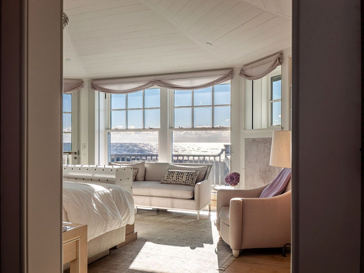 This serene primary bedroom features a light-filled seating area positioned in front of large windows that offer expansive ocean views. The room is decorated in a soft, neutral palette with a plush sofa, a comfortable armchair, and a white bed frame, creating a tranquil and luxurious atmosphere. The perspective is framed through a doorway, emphasizing the inviting transition into this bright, coastal-inspired retreat.