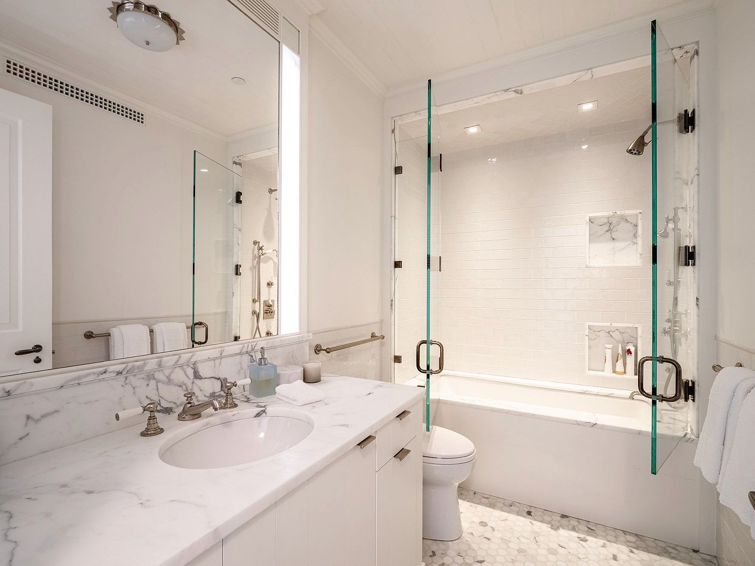 This bright and elegant bathroom features a white vanity topped with a light-veined stone counter and a polished metal faucet. A glass-enclosed shower and tub combination is tiled in white, complemented by a patterned stone floor. The space is illuminated by a large mirror and integrated lighting, creating a clean and sophisticated atmosphere.
