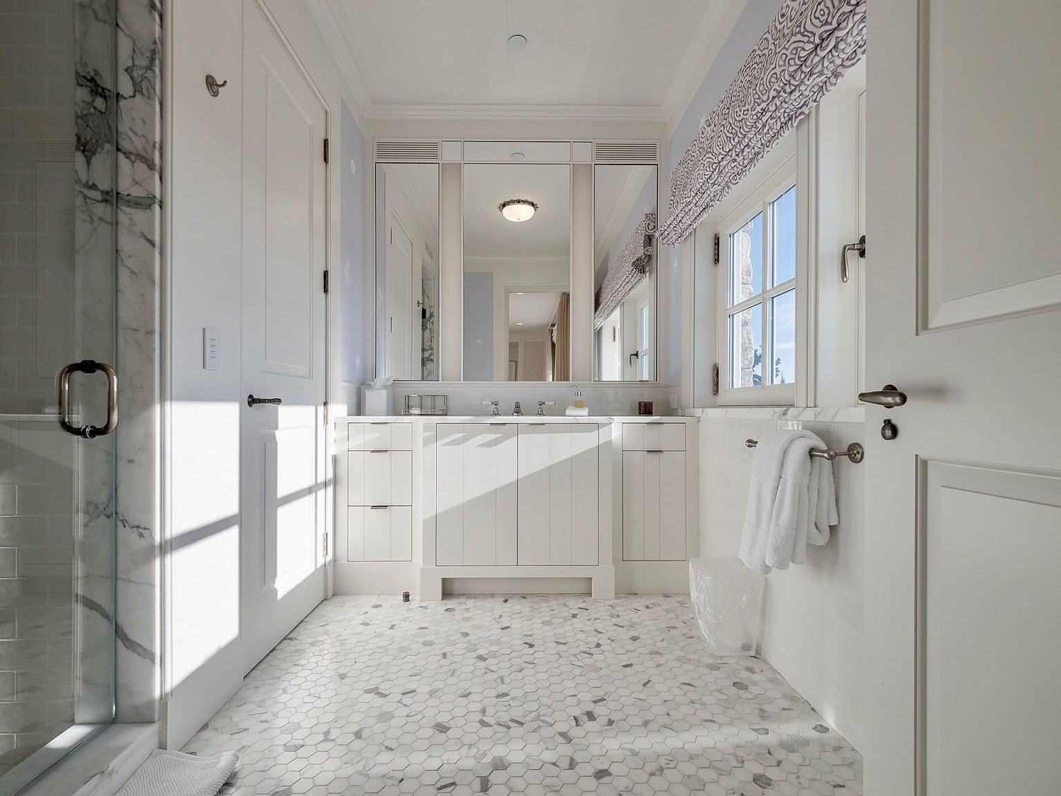 This bright and airy bathroom features a central vanity with a large mirror, flanked by white cabinetry and topped with a stone counter. The floor is covered in a classic hexagonal patterned tile, while a glass-enclosed shower is visible on the left. A window with a patterned valance allows natural light to fill the space, creating a clean and elegant atmosphere.