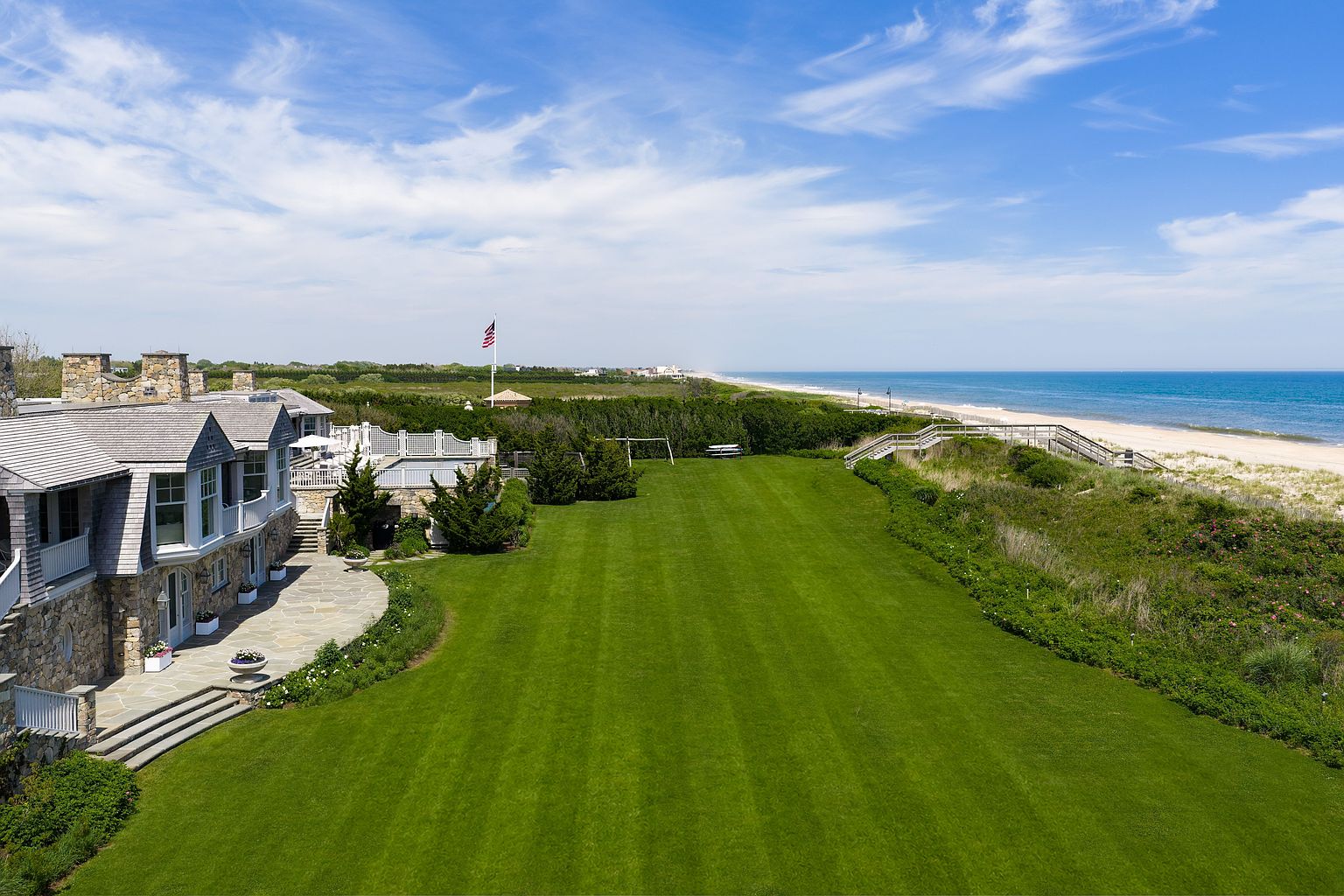 This elevated aerial perspective captures a sprawling coastal estate featuring a stone-clad residence with a shingled roof and a large, manicured lawn. A stone patio extends from the home, leading out to the expansive green space that borders a private wooden staircase descending to the sandy beach. The scene is framed by a bright blue sky with wispy clouds, emphasizing the property's prime oceanfront location and serene atmosphere.