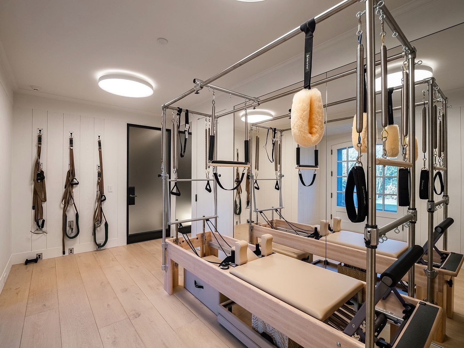 This bright and modern home gym is dedicated to Pilates, featuring a professional-grade reformer machine as the central focus. The room is finished with light wood flooring and white paneled walls, with a large mirror reflecting the equipment to create a sense of depth. Additional exercise straps are neatly mounted on the wall, and the space is illuminated by a sleek, circular flush-mount ceiling light.
