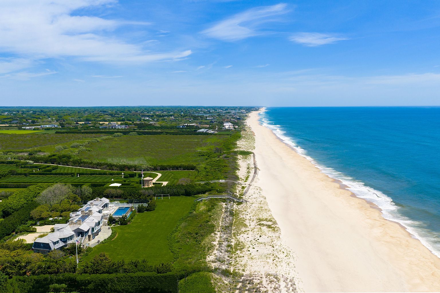 This high-angle aerial perspective captures a sprawling coastal estate situated directly along a pristine, sandy beach. The property features a large, multi-gabled residence with a private swimming pool, expansive manicured lawns, and meticulously maintained hedges. The vast, deep blue ocean stretches to the horizon, contrasting with the lush green landscape and the light-colored dunes that separate the home from the shoreline.