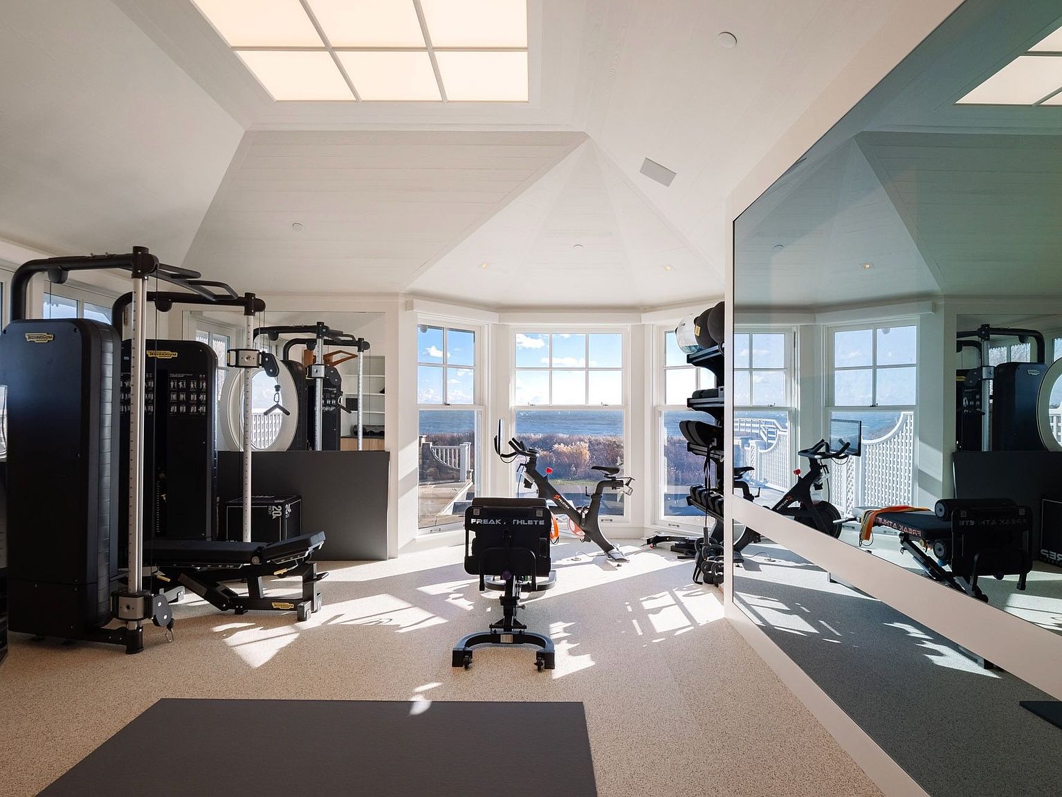 This bright and airy home gym features a professional-grade cable machine, a stationary bike, and a weight bench, all positioned to take advantage of the expansive bay windows overlooking the ocean. The room is topped with a large skylight that floods the space with natural light, while a wall-to-wall mirror on the right enhances the sense of depth and openness. The neutral color palette and clean lines create a sophisticated and motivating environment for fitness.