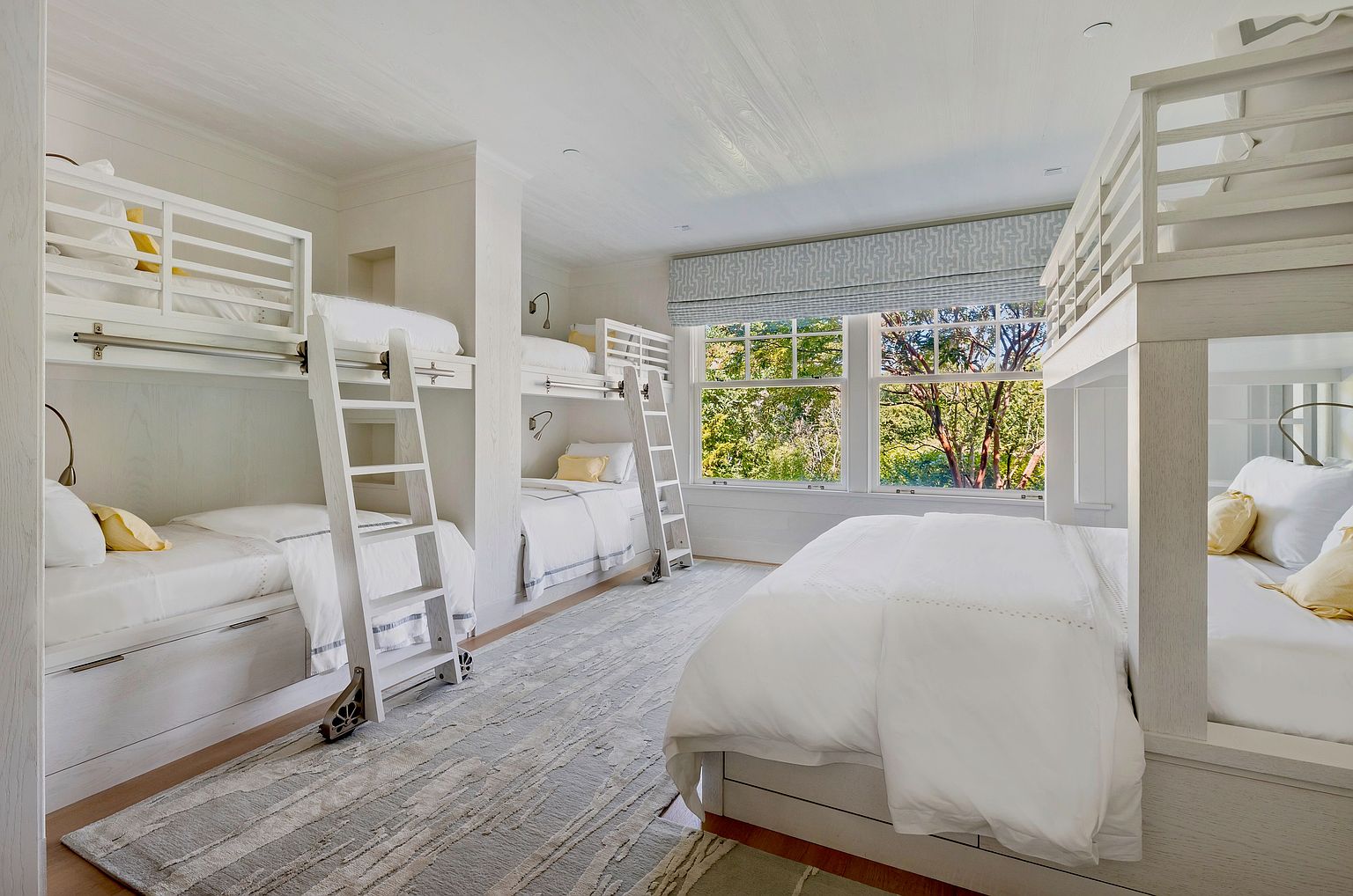 This spacious, light-filled bunk room features multiple white-framed bunk beds arranged to maximize sleeping capacity, making it an ideal space for guests or children. The room is anchored by a large, textured area rug over light wood flooring and is illuminated by natural light streaming through two large windows dressed in patterned roman shades. The clean, neutral aesthetic is accented by soft yellow pillows, creating a bright and inviting atmosphere.