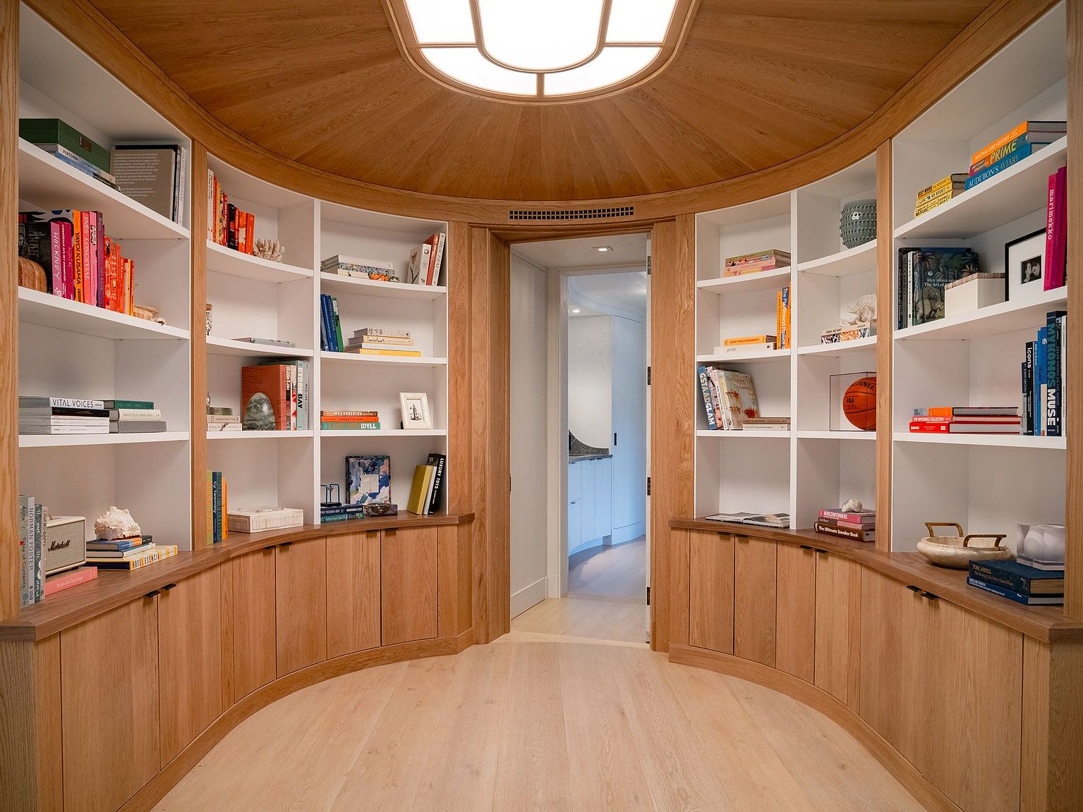 This elegant hallway features a curved design with custom light wood built-in shelving and cabinetry that frame a central doorway. The space is illuminated by a large, circular recessed ceiling light, creating a warm and inviting atmosphere. The light wood flooring and clean, minimalist aesthetic provide a sophisticated transition between rooms.