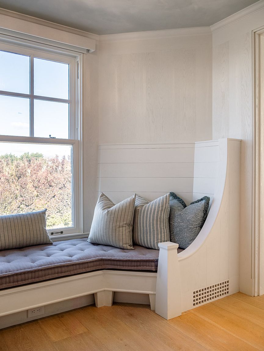 This image highlights a custom-built, L-shaped window seat nestled into a corner, featuring horizontal shiplap paneling and a tufted, neutral-toned cushion. The built-in unit includes a decorative end post and a ventilation grille, complemented by a collection of striped and textured throw pillows. The scene is bathed in natural light from the adjacent window, showcasing a clean, transitional design aesthetic.
