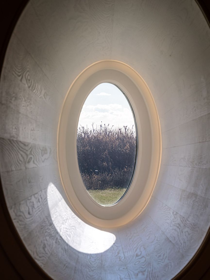 This image captures a unique, oval-shaped window set within a curved, light-toned wood-paneled wall. The perspective is framed from inside, looking out toward a natural landscape of tall grasses and a bright, cloudy sky. The minimalist design emphasizes clean lines and a seamless integration between the interior architecture and the outdoor environment.
