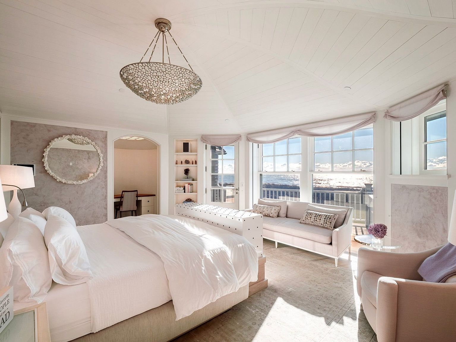 This elegant primary bedroom features a vaulted ceiling with white paneling and a central crystal chandelier, creating a bright and airy atmosphere. A plush bed sits at the center, facing a sofa positioned in front of large windows that offer expansive ocean views. The room includes a soft color palette, a circular wall mirror, and a small adjacent office nook, all contributing to a serene and luxurious coastal aesthetic.