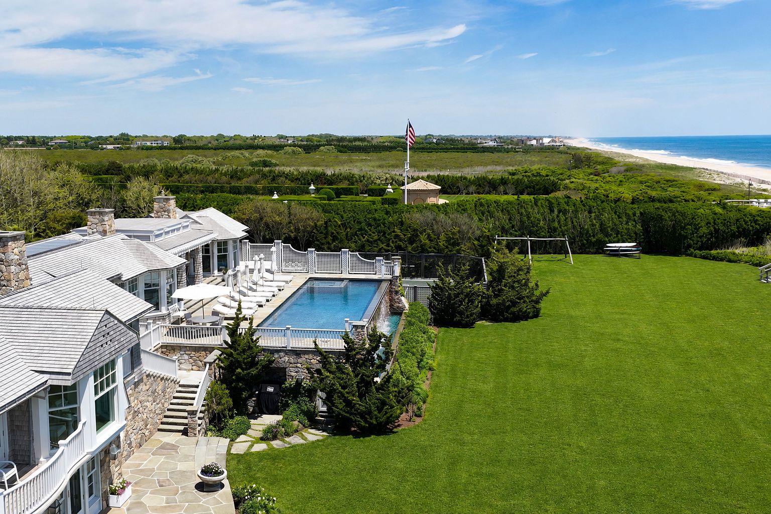 This high-angle aerial view captures a luxurious coastal estate featuring a multi-level shingle-style home with a stone foundation and a prominent infinity-edge pool. The property boasts an expansive, manicured green lawn that transitions into a lush hedge-lined landscape, with the ocean coastline visible in the distance. A tall flagpole stands prominently in the background, emphasizing the grand scale and prime location of this seaside residence.