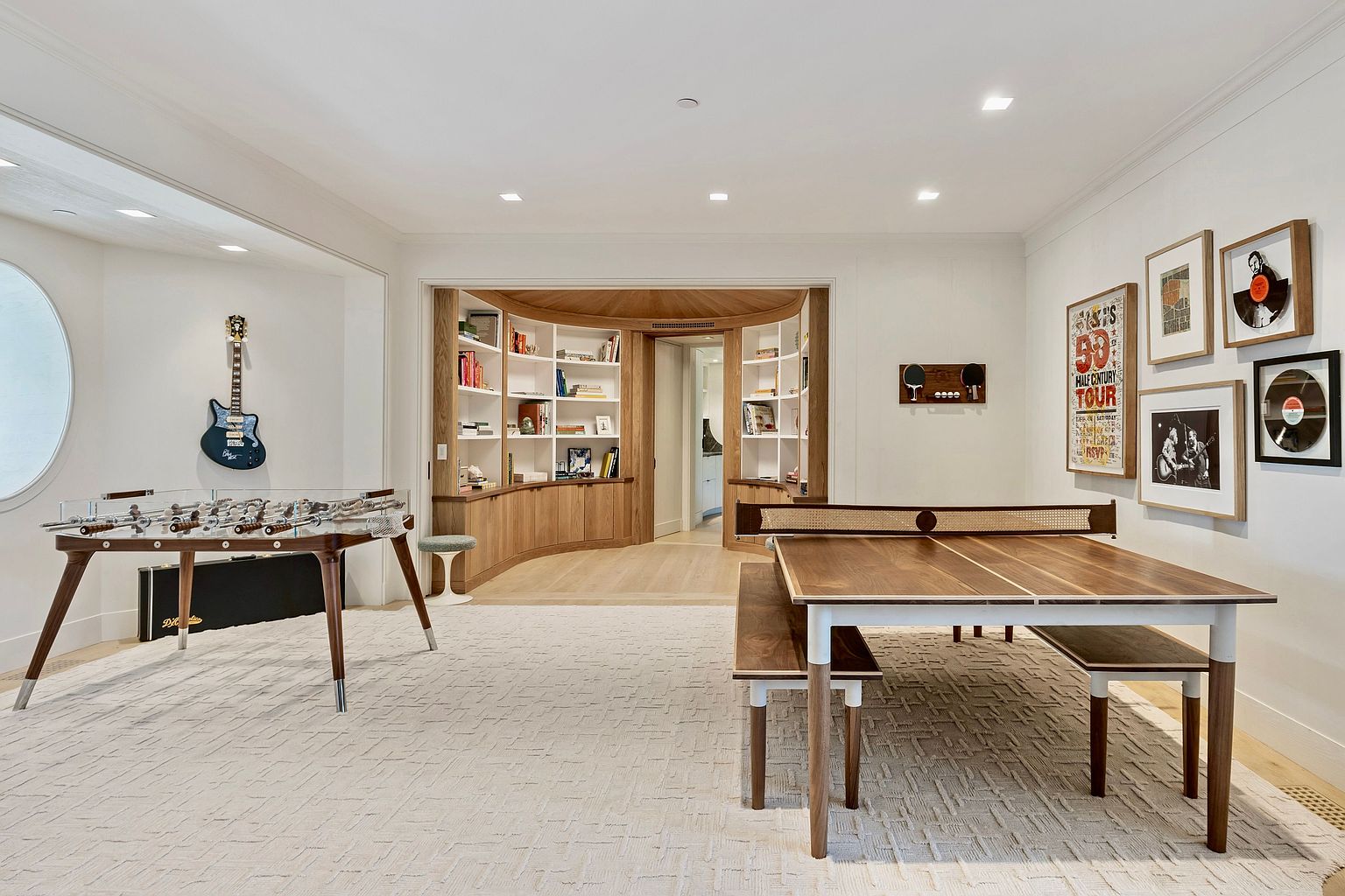 This spacious game room features a modern foosball table on the left and a versatile table tennis setup with matching benches on the right. A curved, built-in shelving unit with light wood cabinetry serves as a focal point in the background, leading to an adjacent room. The room is decorated with framed music-themed artwork and a wall-mounted guitar, creating a sophisticated yet playful entertainment space.