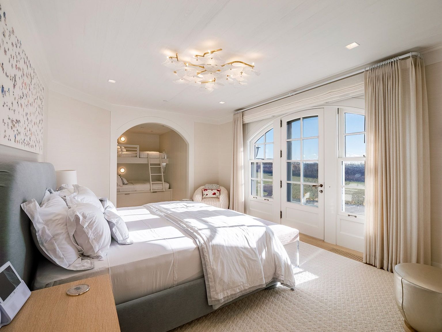 This bright and airy primary bedroom features a large, plush bed with a grey upholstered headboard and crisp white bedding. To the side, a recessed alcove houses built-in bunk beds, while a set of arched glass doors with tall windows allows for abundant natural light and views of the outdoors. The room is accented by a unique, organic-shaped chandelier and a soft, neutral color palette that creates a serene and inviting atmosphere.