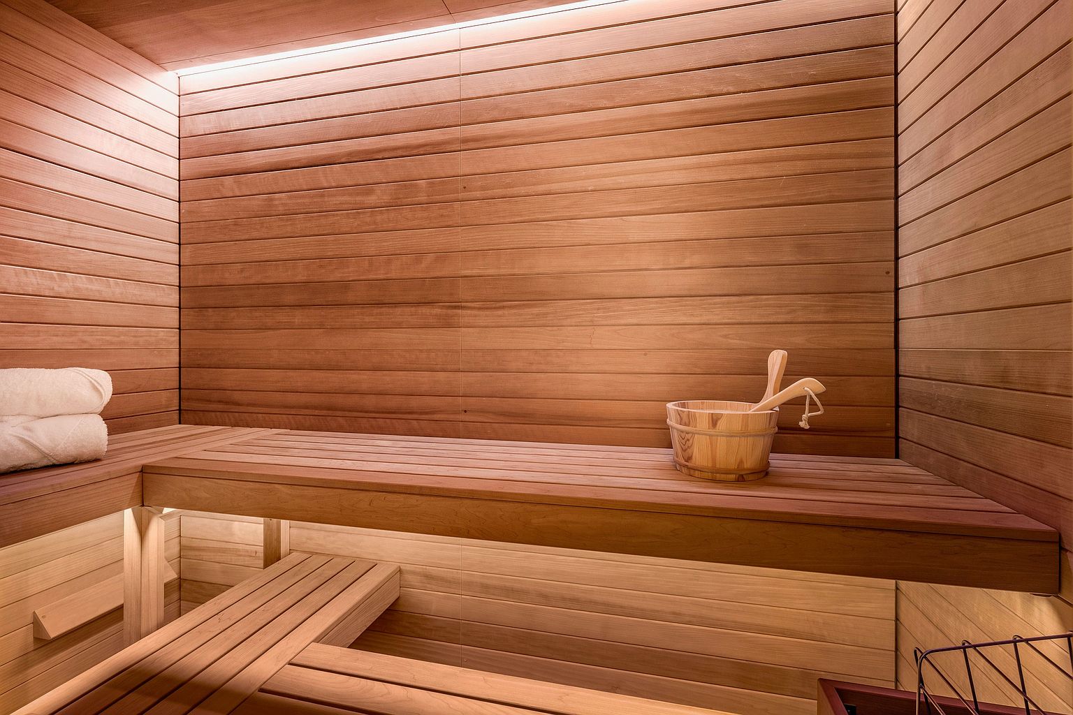 This image showcases a sauna interior, characterized by its warm, light-toned wood paneling on the walls and tiered bench seating. A wooden bucket with a ladle sits on the upper bench, while a stack of white towels rests on the left, creating a relaxing and spa-like atmosphere. The space is illuminated by soft, integrated lighting beneath the benches, enhancing the cozy and inviting aesthetic.