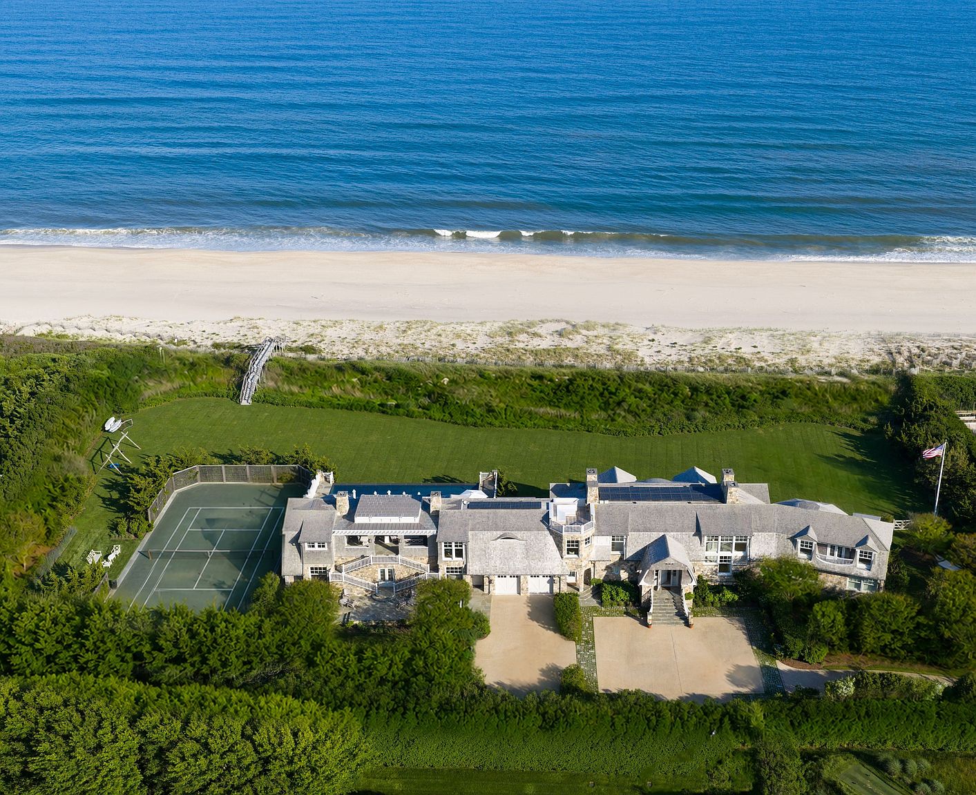 This expansive aerial view captures a luxurious beachfront estate featuring a sprawling shingle-style residence with multiple rooflines and solar panels. The property includes a private tennis court, a manicured lawn, and a direct wooden walkway leading from the backyard to the sandy beach and ocean. The perspective highlights the home's prime coastal location, emphasizing its seamless integration with the surrounding natural landscape.