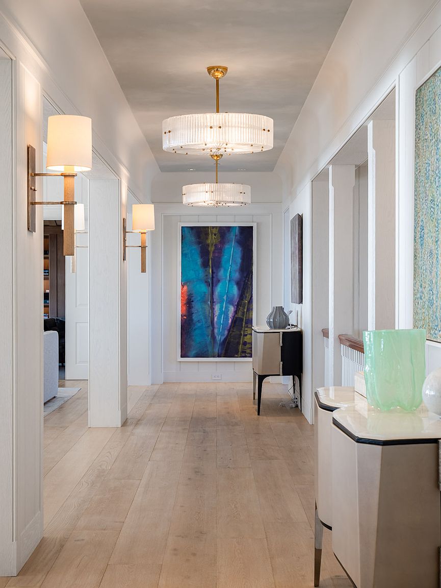 This elegant, light-filled hallway features light wood flooring and crisp white walls, creating a sophisticated and airy atmosphere. The space is punctuated by two tiered, ribbed-glass chandeliers and matching wall sconces, leading the eye toward a vibrant, large-scale abstract painting at the end of the corridor. A sleek console table with a decorative glass vase adds a refined touch to the transition space, which also offers a glimpse into an adjacent room.