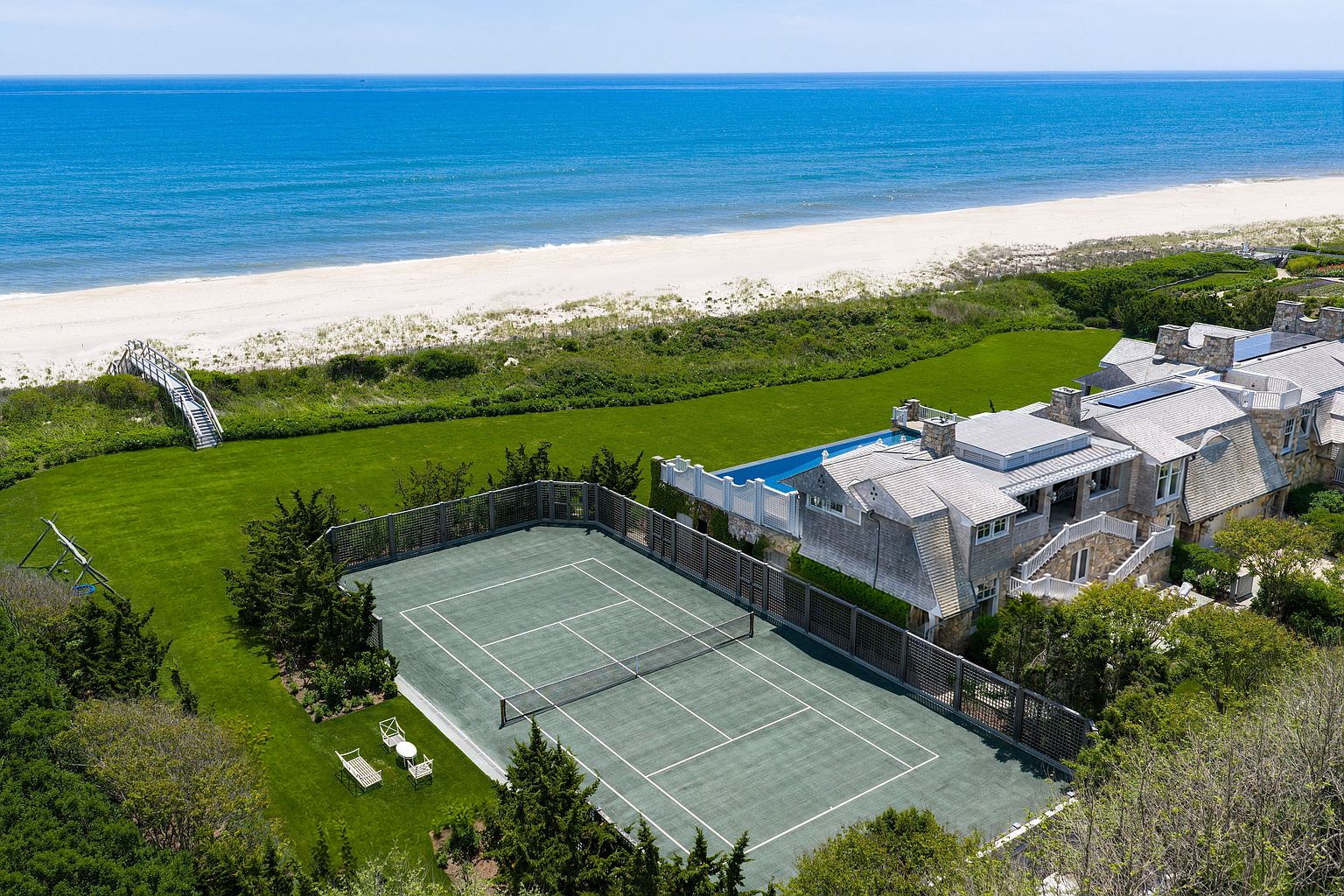 This high-angle aerial perspective captures a sprawling coastal estate featuring a private tennis court, a lush green lawn, and a swimming pool overlooking the ocean. The shingled residence showcases classic architectural details, including multiple stone chimneys and a multi-level roofline, set against the backdrop of a pristine sandy beach and the Atlantic horizon. A private wooden walkway provides direct access from the property to the dunes and shoreline, emphasizing the home's exclusive waterfront location.