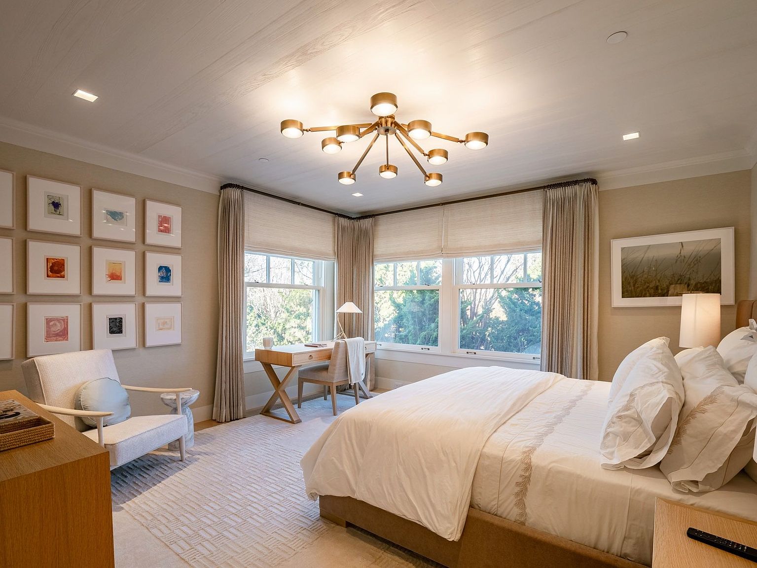 This serene primary bedroom features a neutral color palette, a plush bed with white linens, and a dedicated workspace tucked into the corner by the windows. A gallery wall of framed artwork adds a personal touch, while a modern multi-arm chandelier serves as a striking focal point. The room is bathed in natural light from large windows dressed in soft drapery, creating a calm and inviting atmosphere.
