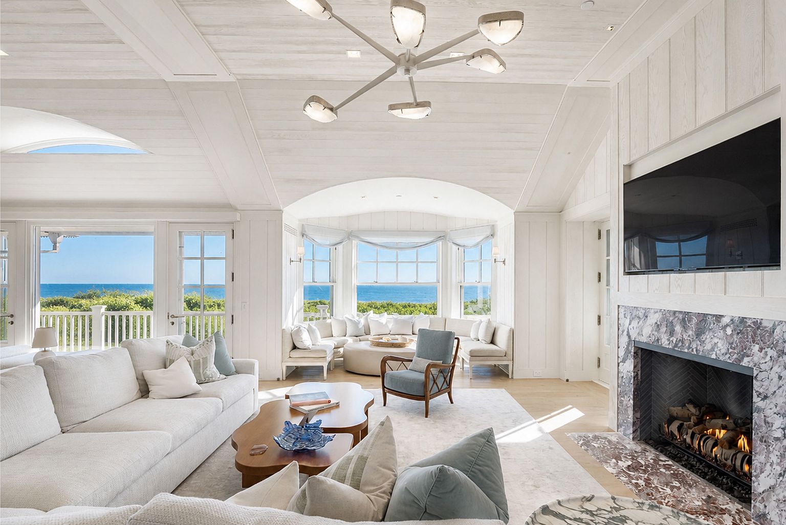 This bright and airy living room features a coastal aesthetic with light wood-paneled ceilings and walls, centered around a stone-clad chimney breast with a built-in television. A large sectional sofa and a blue accent chair are arranged around a unique, multi-tiered wooden coffee table, while a curved window nook offers panoramic ocean views. The space is illuminated by a modern, multi-armed chandelier and abundant natural light, creating a serene and luxurious atmosphere.
