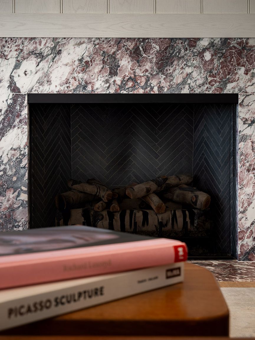 This close-up shot highlights a fireplace with a striking, multi-toned stone surround featuring deep red and dark grey veining. The firebox is lined with dark, herringbone-patterned tile and contains a stack of birch logs, while two art books rest on a wooden surface in the foreground. The composition creates a sophisticated, textured focal point that emphasizes high-end interior finishes.