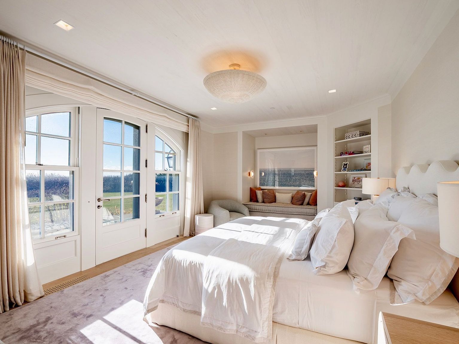 This bright and airy primary bedroom features a plush king-sized bed as the centerpiece, complemented by a soft, light-toned rug and a textured ceiling. A cozy window seat nook is built into the wall, accented by warm-toned throw pillows and flanked by open shelving. Large glass doors with arched transoms provide abundant natural light and a seamless connection to the outdoor landscape.
