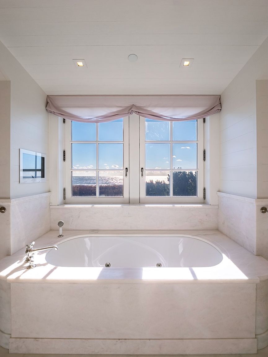 This serene primary bathroom features a deep soaking tub encased in a light-toned stone surround, positioned directly beneath a pair of large, multi-pane windows that frame a scenic ocean view. The space is finished with clean, horizontal wall paneling and soft, draped window treatments, creating a bright and tranquil atmosphere. The symmetrical composition highlights the tub as the central focal point, emphasizing a luxurious and relaxing coastal aesthetic.