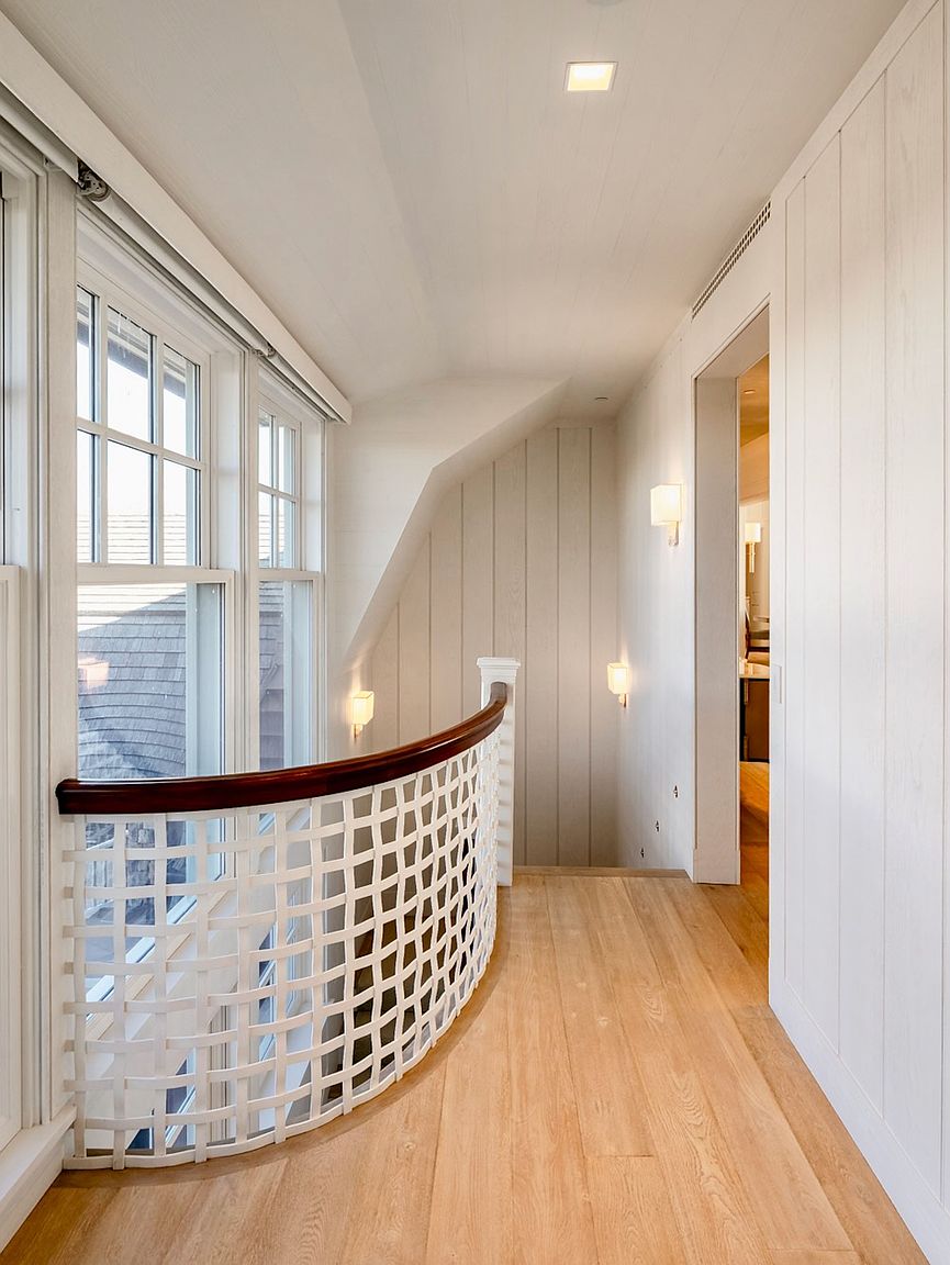 This bright hallway features light wood flooring and white vertical wall paneling, creating a clean and airy aesthetic. A curved staircase railing with a decorative white lattice pattern and a dark wood handrail overlooks the lower level, while wall-mounted sconces provide warm accent lighting. The perspective looks down the corridor toward an open doorway, highlighting the architectural transition between spaces.