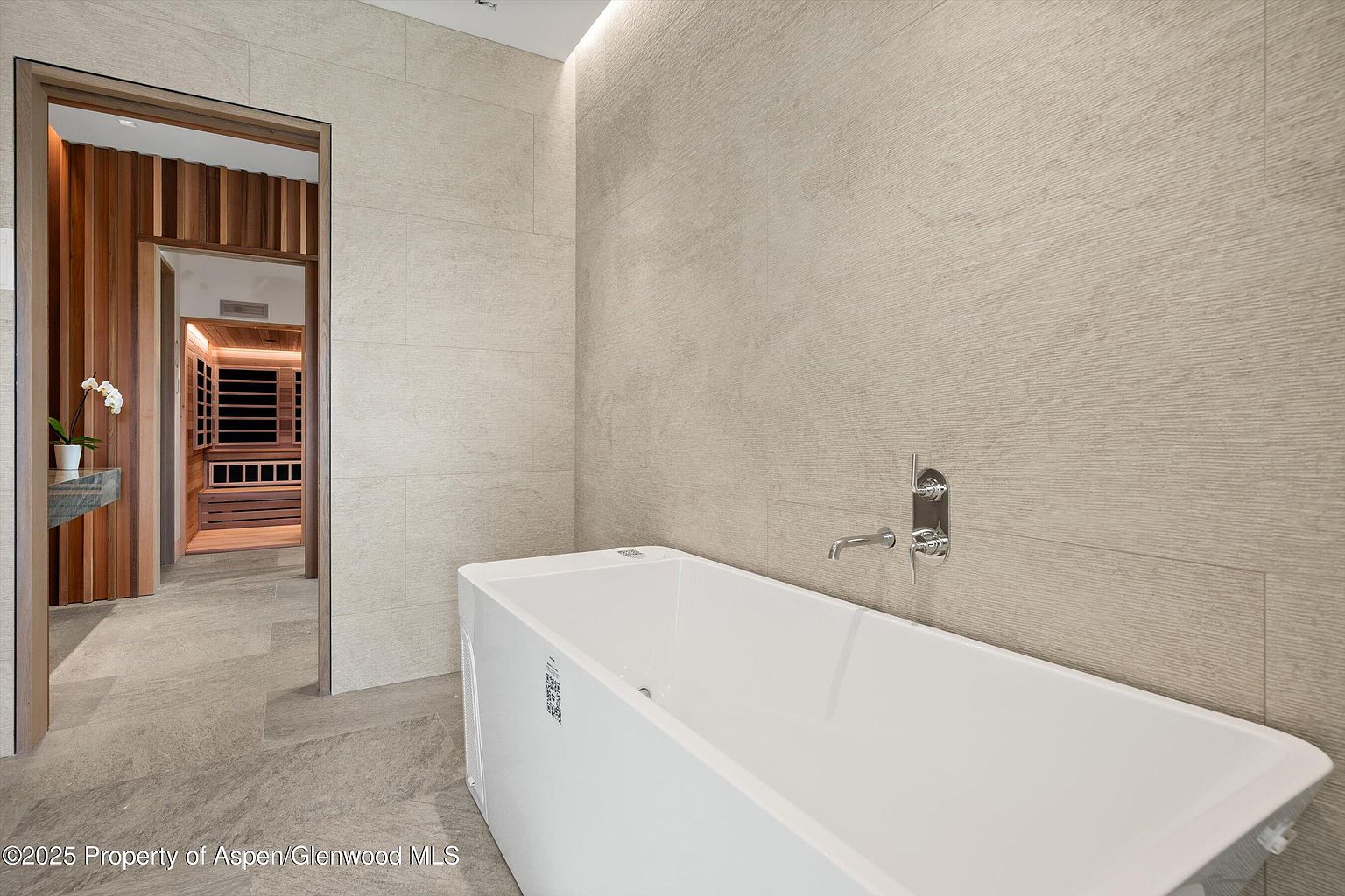 This is a modern primary bathroom featuring a sleek, freestanding white bathtub with chrome fixtures. The walls are covered in light beige textured tiles, and the floor is a matching stone. An open doorway reveals a wood-paneled sauna, adding a luxurious touch to the space.