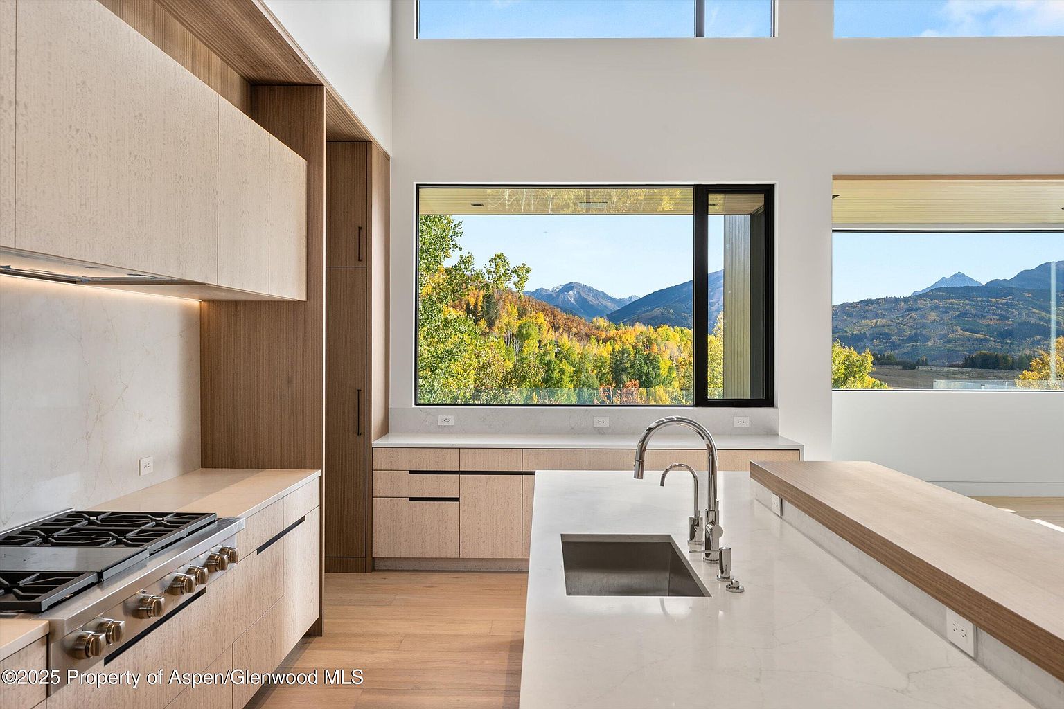 This is a bright, modern kitchen featuring light wood cabinetry and a large island with a white countertop and a built-in sink. Large windows offer stunning views of the surrounding mountains and trees, bringing natural light into the space. The kitchen is equipped with stainless steel appliances and has a clean, minimalist design.
