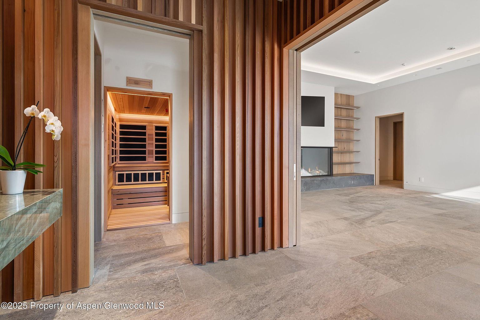 This interior shot showcases a modern hallway with a sauna visible through one doorway and a living area through another. The hallway features wood slat walls and neutral-toned tile flooring, creating a warm and inviting atmosphere. The design emphasizes clean lines and natural materials, contributing to a luxurious and contemporary aesthetic.