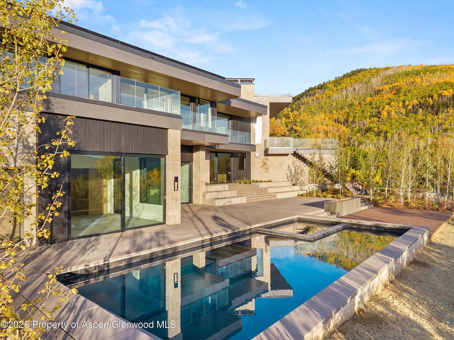 This image showcases the exterior of a modern home, focusing on the pool and spa area. The pool features clean lines and is surrounded by a stone patio, with a wooden deck adjacent to it. The house has large windows and balconies, offering views of the surrounding landscape, which includes a hillside covered in autumn foliage.