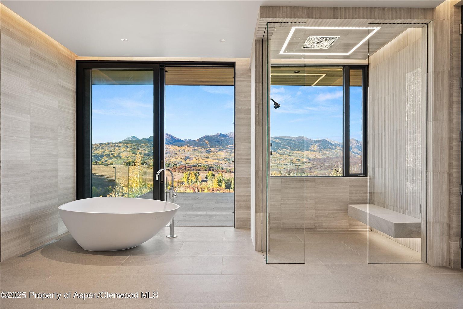 This is a luxurious primary bathroom featuring a freestanding white bathtub positioned in front of a large sliding glass door that offers stunning mountain views. Adjacent to the tub is a modern glass-enclosed shower with a built-in bench and a rainfall showerhead. The bathroom is finished with neutral-toned tile and sleek fixtures, creating a spa-like atmosphere.