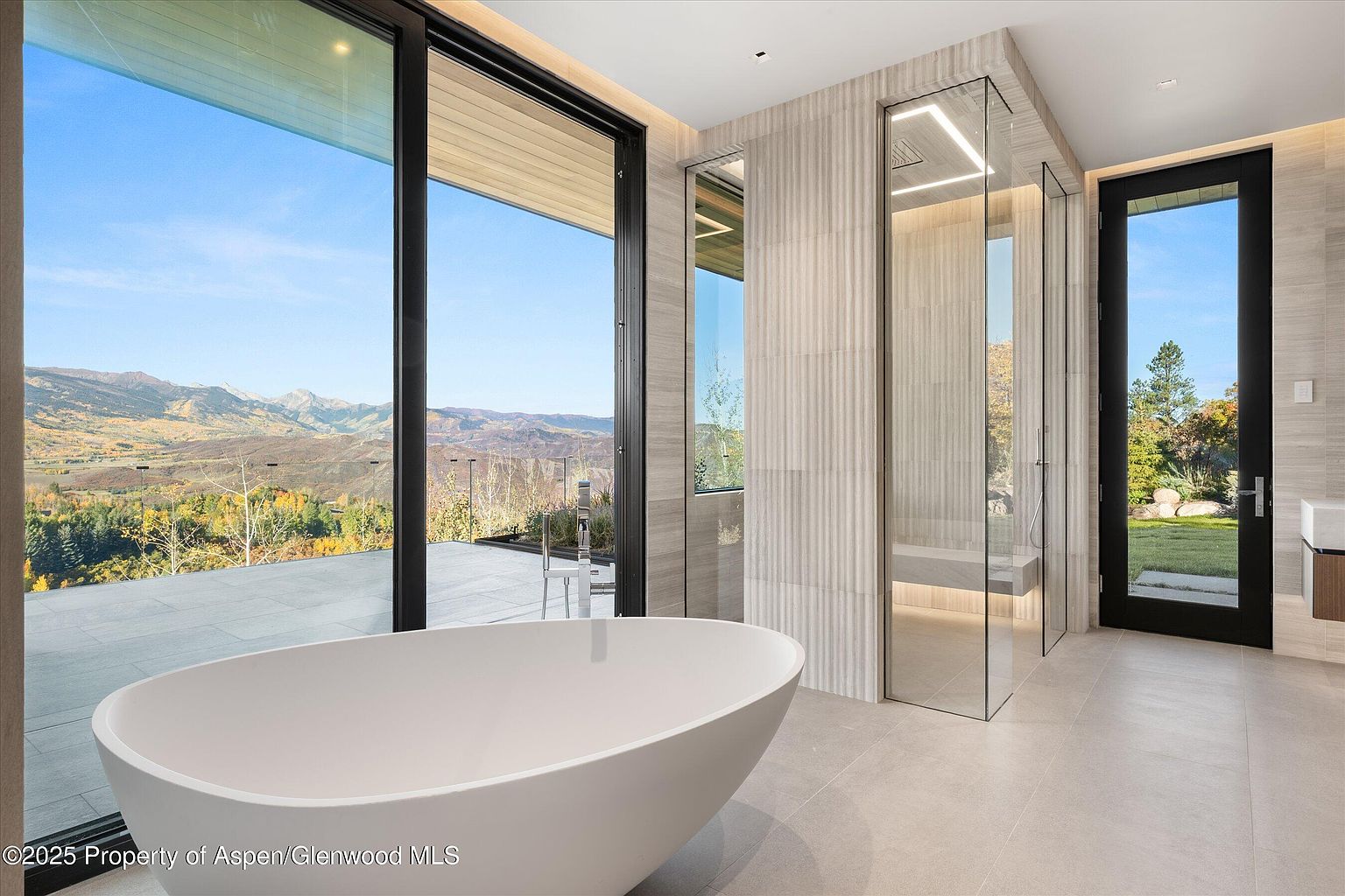 This is a luxurious primary bathroom featuring a freestanding oval bathtub positioned to take advantage of a large window with a scenic mountain view. The bathroom includes a glass-enclosed shower and a door leading to the outside, enhancing the connection with nature. The neutral color palette and clean lines create a spa-like atmosphere.
