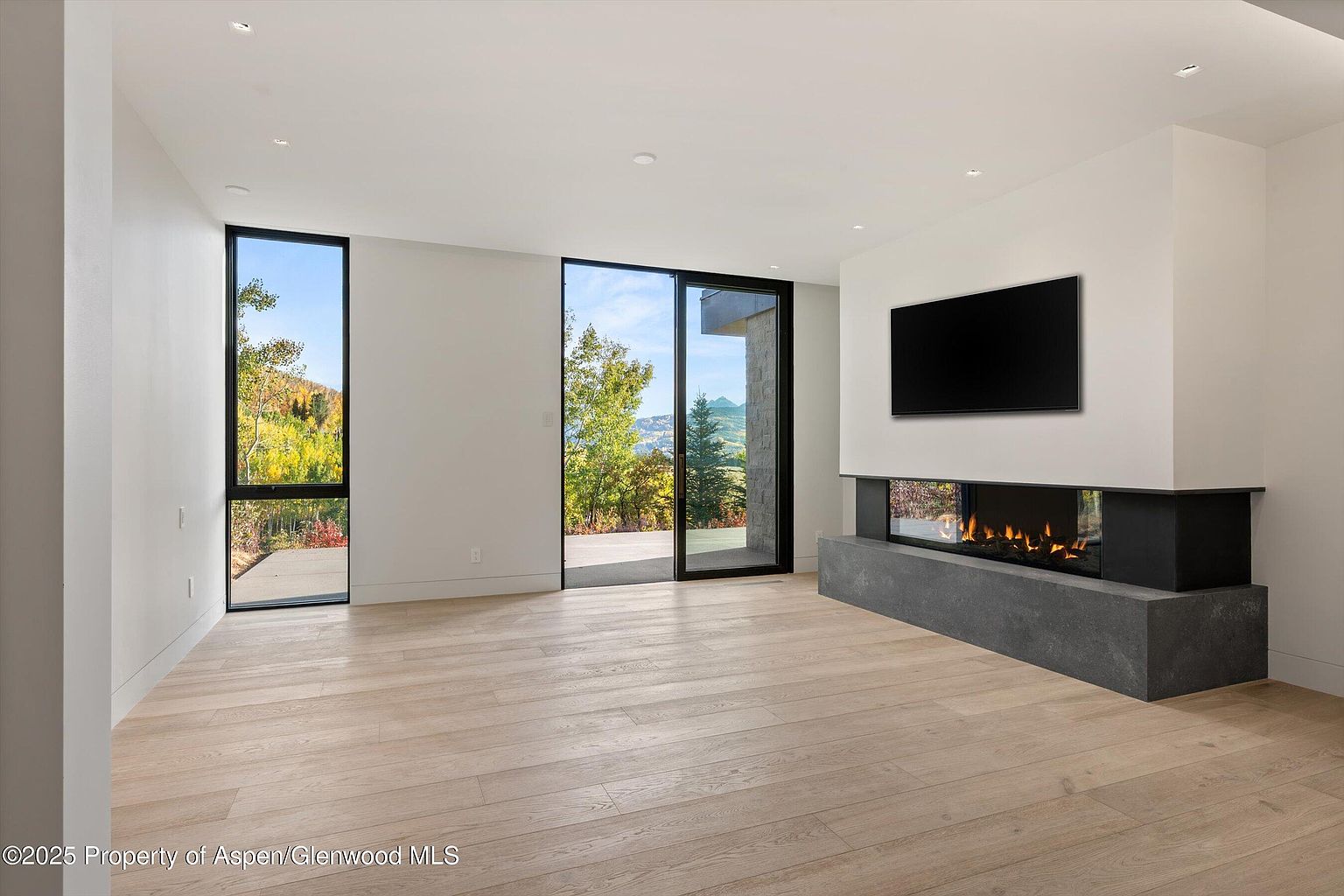 This is a bright and modern living room featuring light wood flooring, white walls, and large windows offering scenic views. A sleek, contemporary fireplace with a mounted television above it serves as a focal point. The room exudes a minimalist and luxurious feel, perfect for relaxation and entertainment.