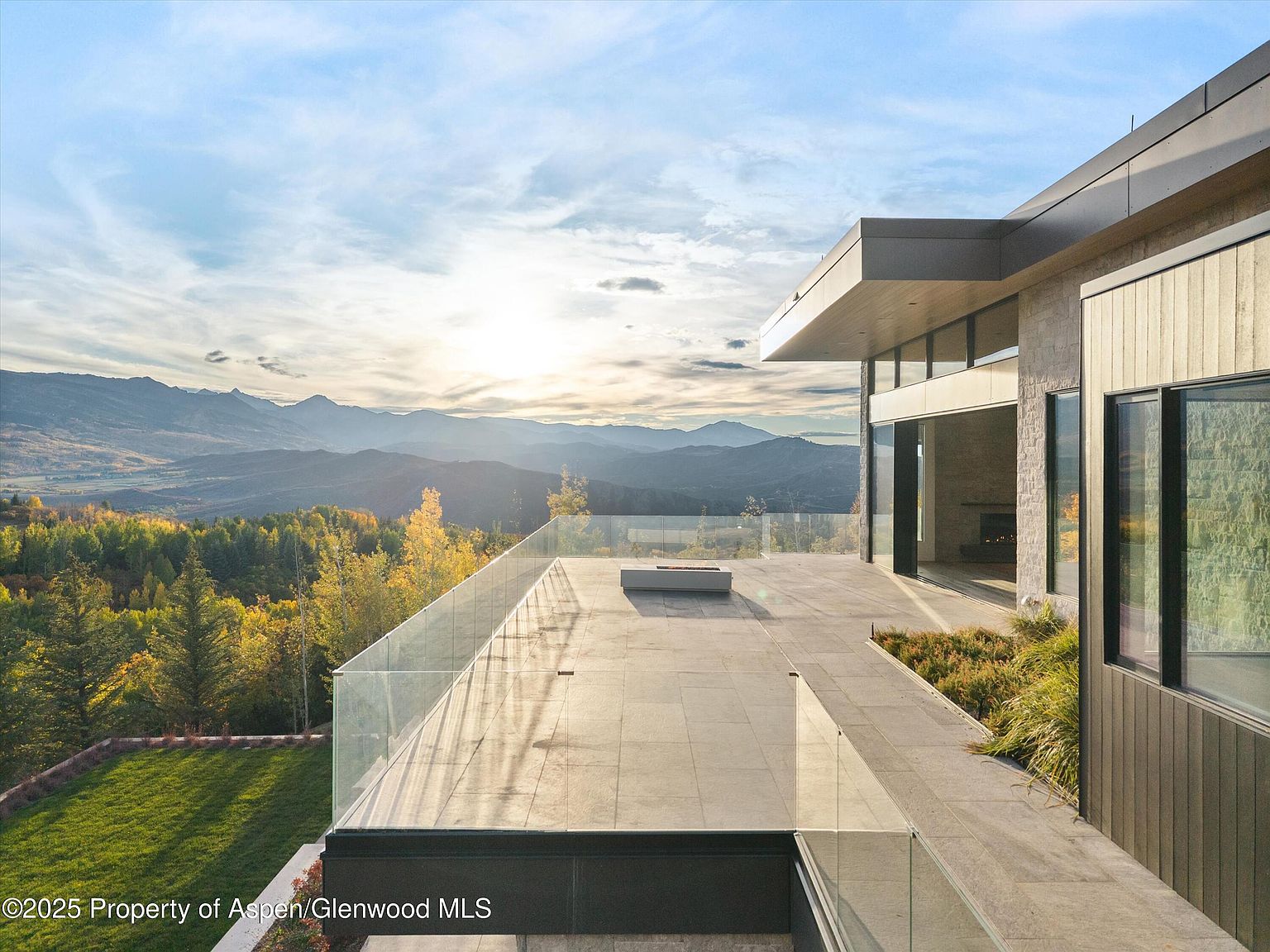 This image showcases a modern home's expansive patio/deck area with glass railings, offering unobstructed views of a stunning mountain landscape at sunset. The architectural design is sleek and contemporary, featuring clean lines and a combination of stone and metal elements. The outdoor space is perfect for entertaining or relaxing while enjoying the breathtaking scenery.