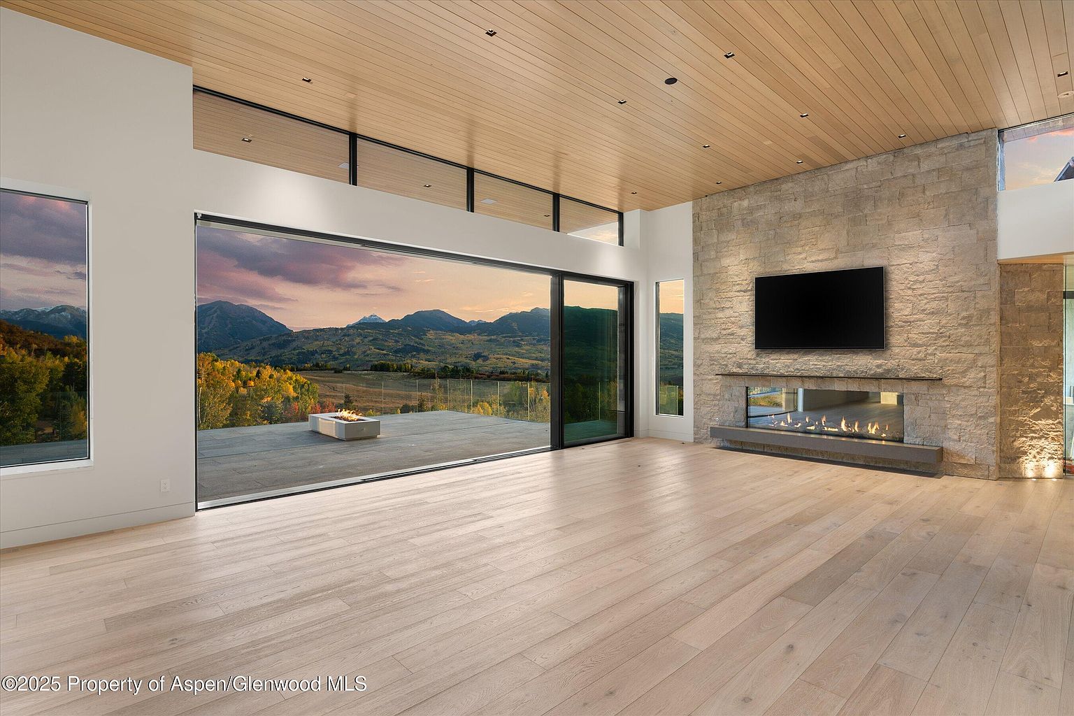This is a spacious living room featuring a modern design with a stone accent wall and a linear fireplace beneath a large television. Expansive sliding glass doors offer panoramic views of the mountains and lead to an outdoor patio. The room is bathed in natural light, highlighting the light wood flooring and creating a warm, inviting atmosphere.