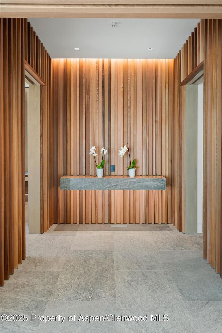 This interior shot showcases a modern hallway with striking wood paneling that creates a warm and inviting atmosphere. A floating stone shelf adorned with orchids adds a touch of elegance, while the neutral-toned tile flooring complements the natural wood tones. The hallway is well-lit, highlighting the architectural details and creating a sense of spaciousness.