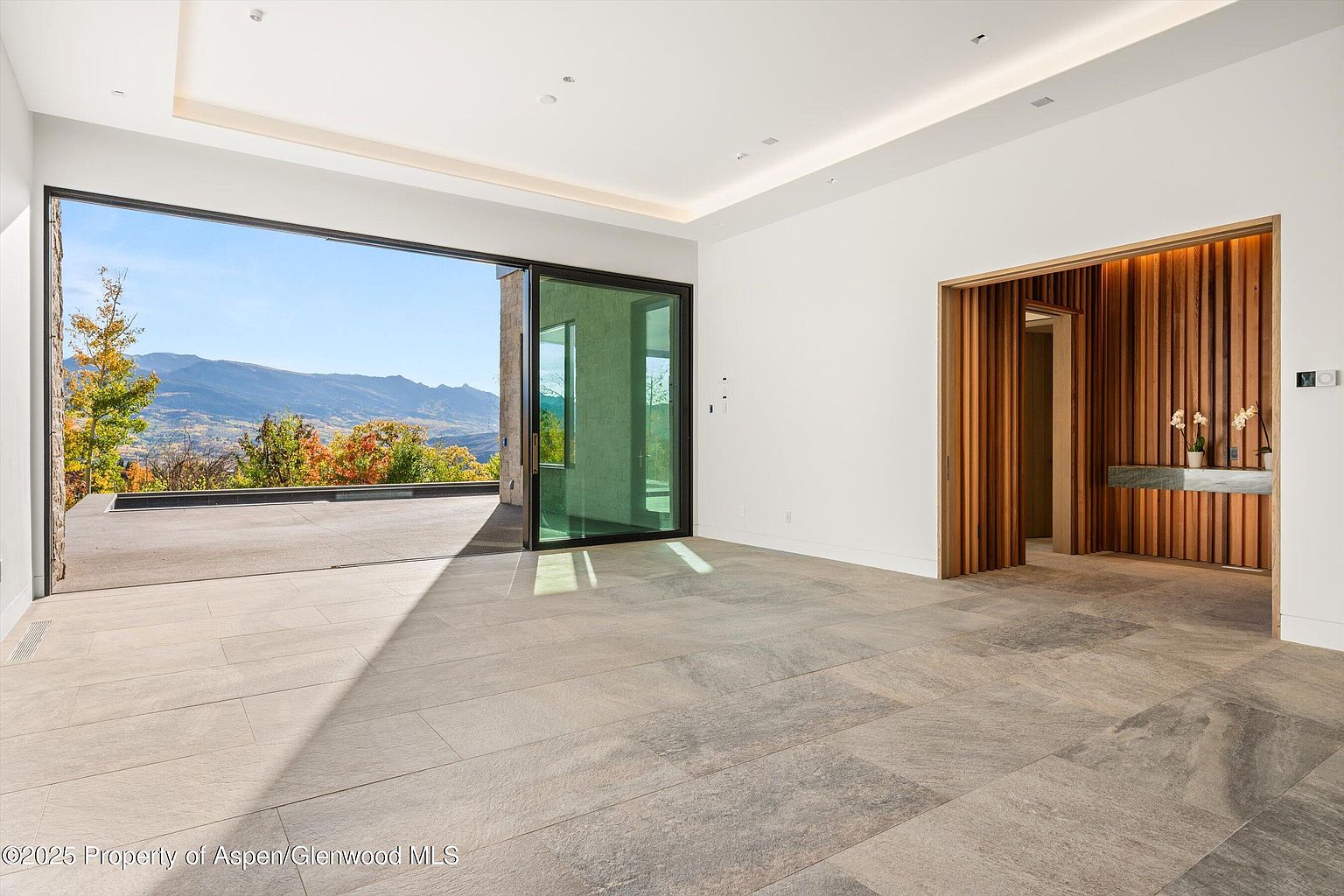This is a spacious living room with large sliding glass doors that open to an outdoor patio and offer stunning mountain views. The interior features a modern design with light-colored tile flooring, white walls, and a unique wooden accent wall with a built-in shelf. The room is bright and airy, creating a seamless transition between indoor and outdoor living spaces.
