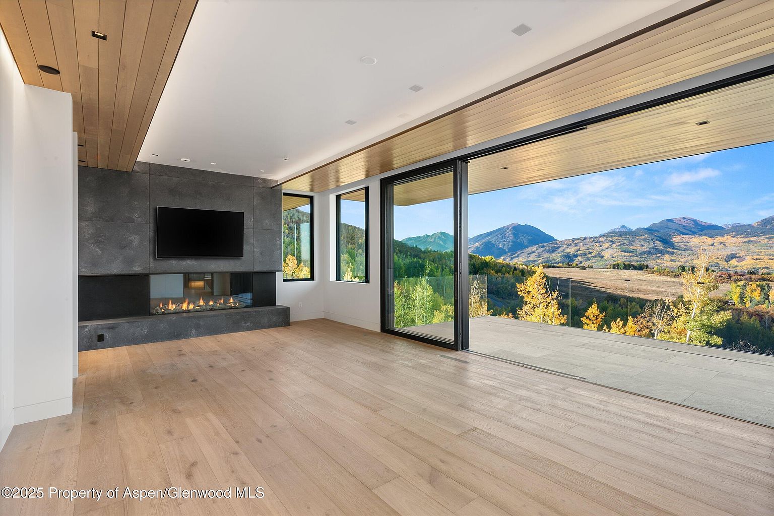 This is a bright and modern living room featuring a sleek fireplace with a large television mounted above. The room has light wood flooring and a wood-paneled ceiling detail, adding warmth to the contemporary design. Large sliding glass doors open to a balcony with stunning mountain views, creating a seamless indoor-outdoor living experience.