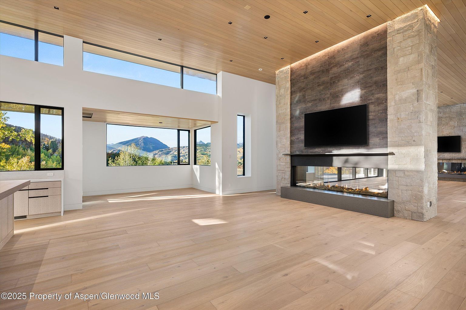 This is a spacious living room featuring a modern fireplace with a large television mounted above. The room has large windows offering scenic mountain views, complemented by light wood flooring and a wood-paneled ceiling. The overall design is contemporary and luxurious, emphasizing natural light and open space.