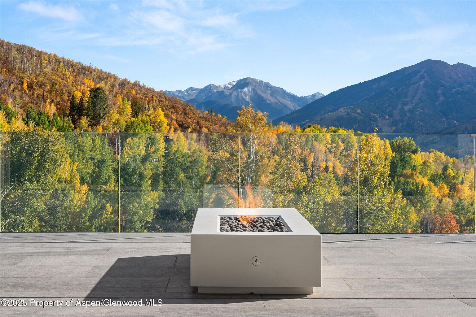 This image showcases a luxurious outdoor patio or balcony with a modern fire pit as the focal point. The patio is paved with large, gray tiles, and a glass railing provides an unobstructed view of the stunning mountain landscape with colorful autumn foliage. The fire pit is a square, light-colored structure filled with dark stones, adding warmth and ambiance to the space.
