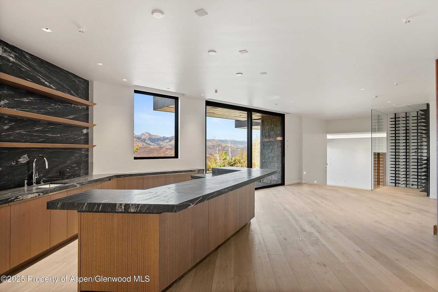 This interior shot showcases a modern kitchen with a unique black marble backsplash and open shelving. The kitchen island features a dark countertop and wood paneling, complementing the light wood flooring. Large windows offer scenic views, and a glass-enclosed wine cellar is visible in the background, adding a touch of luxury.