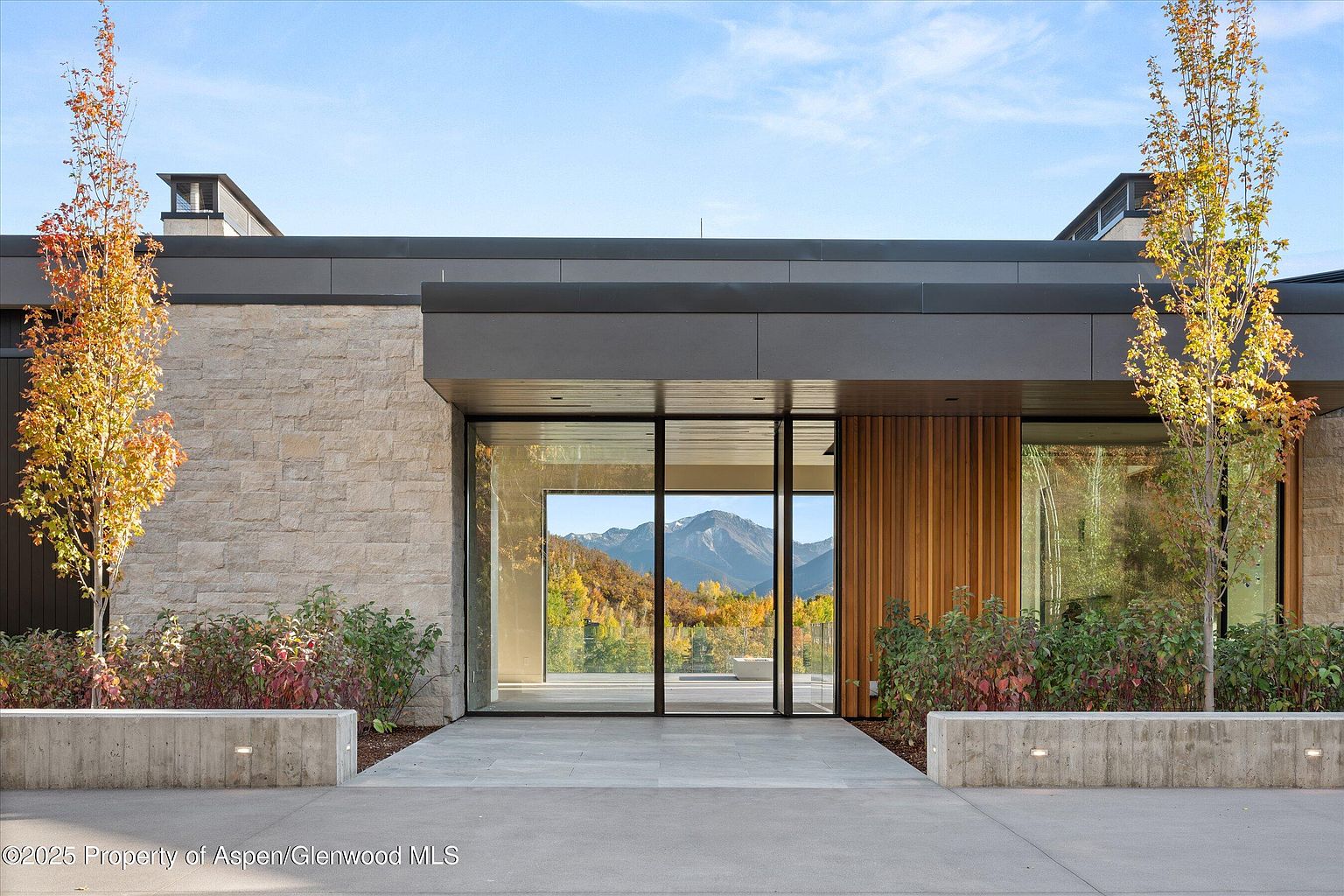 This is a striking front entryway of a modern home, featuring a combination of stone and wood paneling. Large glass windows offer a glimpse of the mountain view beyond, creating a seamless connection between the interior and exterior. The entryway is framed by manicured landscaping and concrete planters, adding to the property's curb appeal.