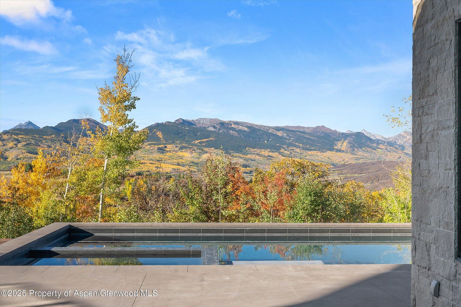 This image showcases a luxurious pool and spa area with a stunning mountain backdrop. The pool's dark tile contrasts beautifully with the light-colored stone patio, creating a modern and elegant aesthetic. The view of the mountains and fall foliage adds a sense of serenity and exclusivity, making it an ideal outdoor space for relaxation and entertainment.