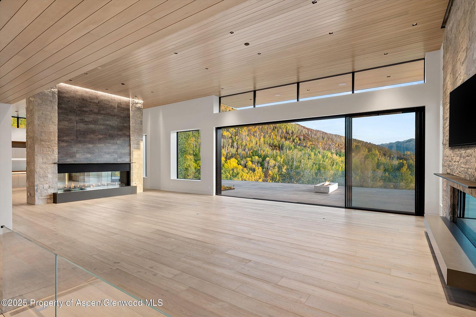 This is a spacious living room featuring a modern design with a stone fireplace and large sliding glass doors that offer a panoramic view of a mountain landscape with autumn foliage. The room has light wood flooring and a wood-paneled ceiling, creating a warm and inviting atmosphere. The open floor plan and natural light enhance the sense of space and luxury.