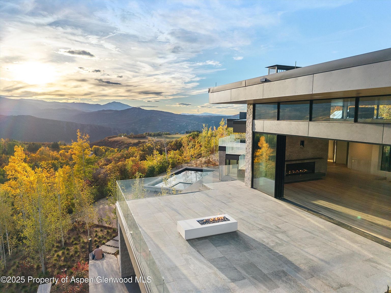 This image showcases a luxurious patio/deck area of a modern home, offering stunning panoramic views of the surrounding mountains and landscape. The patio features a sleek fire pit, glass railings, and seamless access to the interior through large sliding glass doors. The design emphasizes indoor-outdoor living and highlights the property's prime location and high-end finishes.